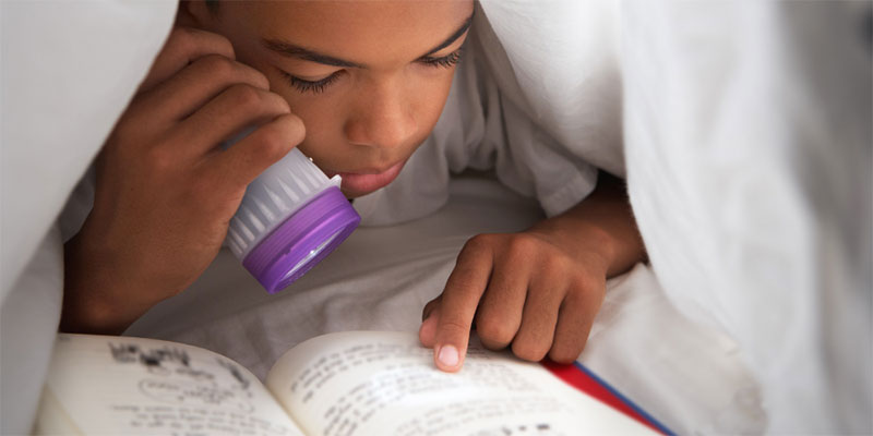 A boy reading a book under the covers with a flashlight